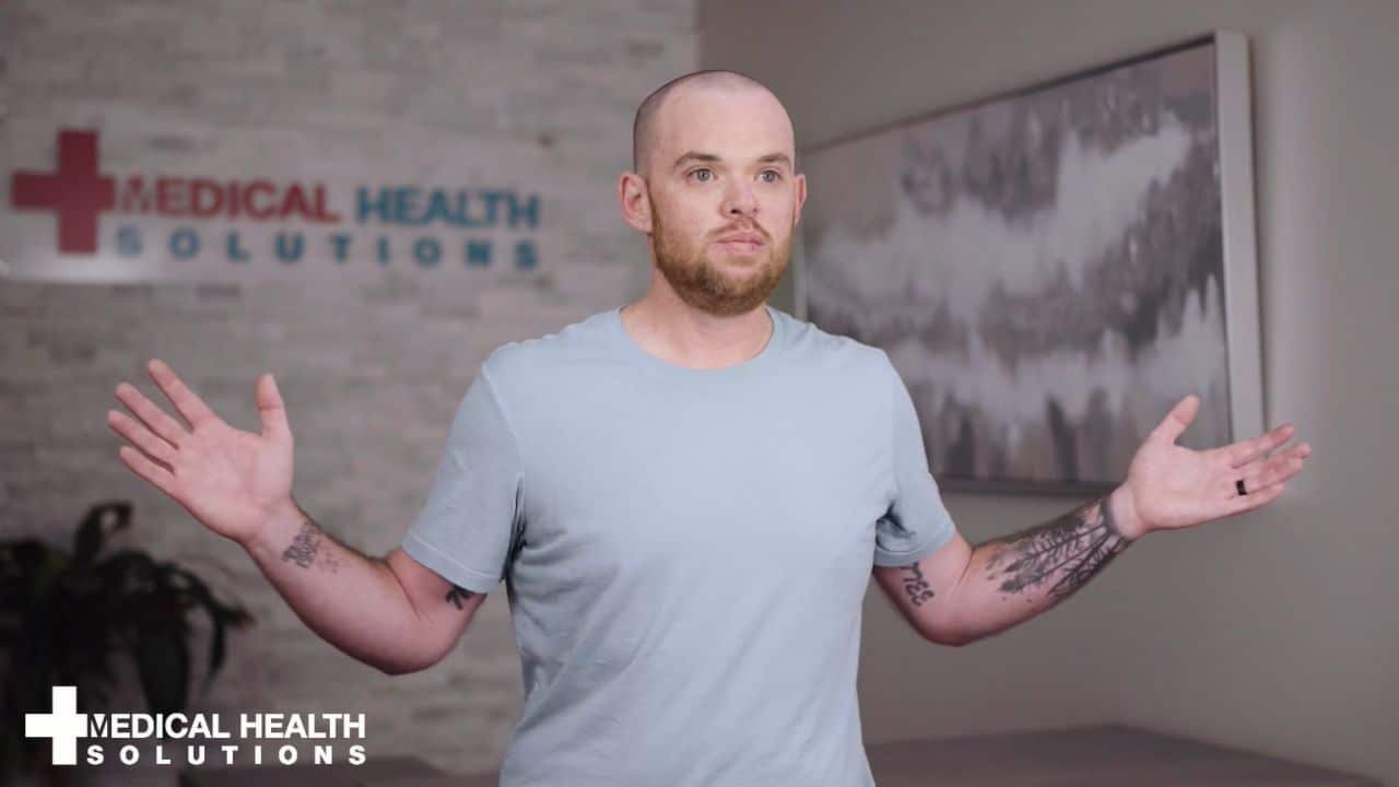 Jeff is standing in front of the wall inside the clinic. The logo is behind him on the wall and his hands arms are spread as he atalk about how he and hos wife Courtney have lost weight on this weight loss program.